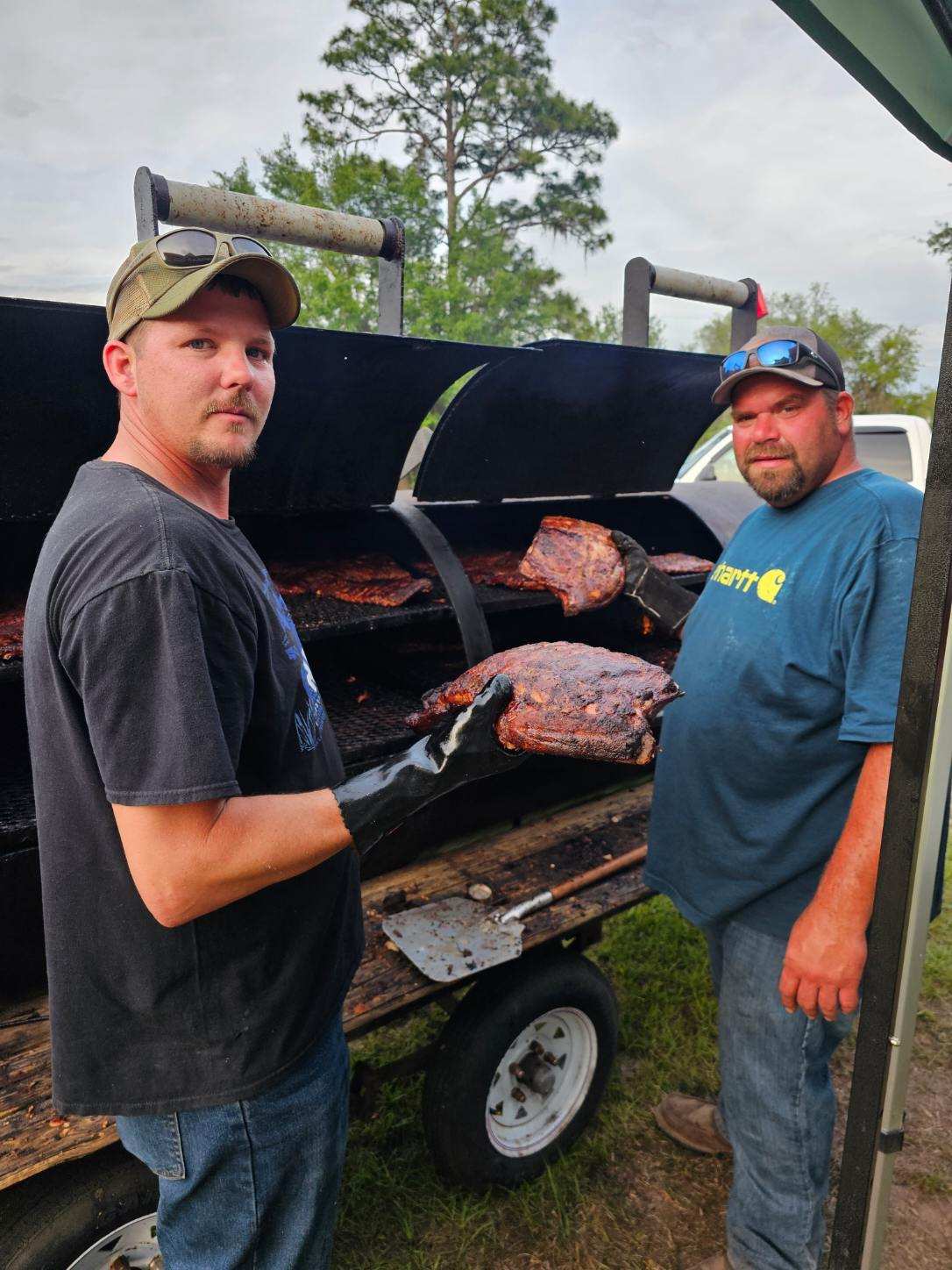 Two men standing in front of a grill with meat on it. 